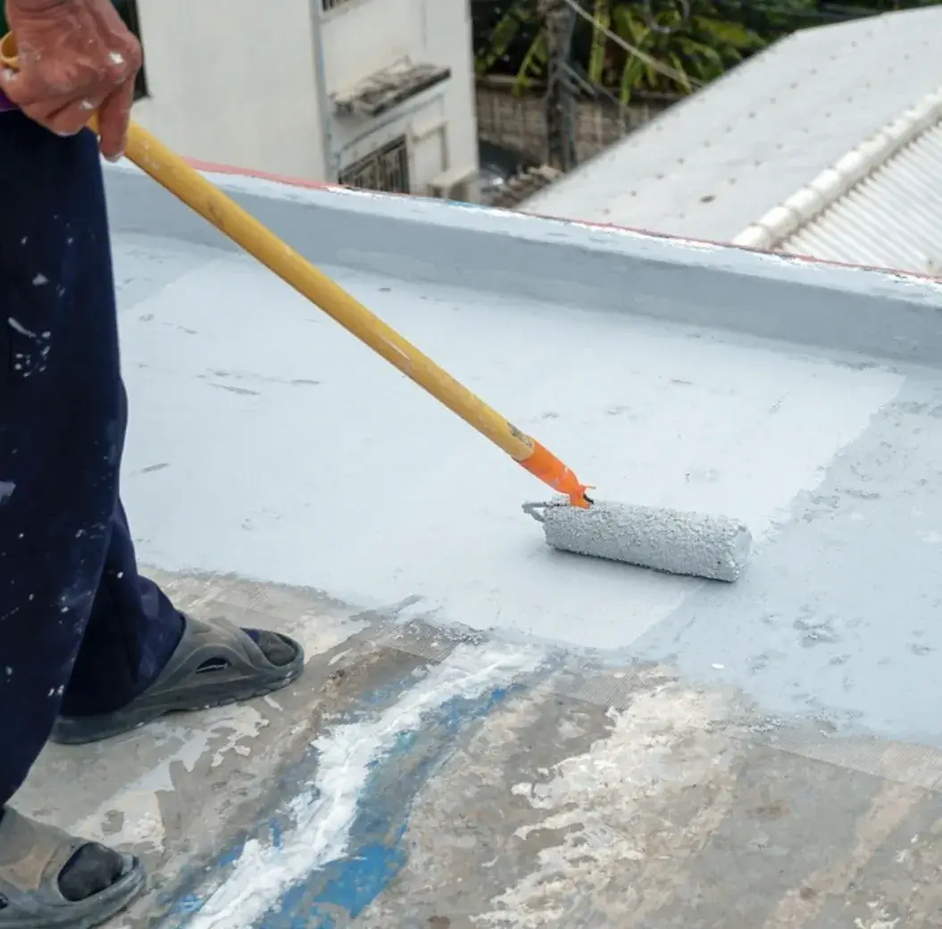 Worker applying waterproofing paint on a flat concrete roof using a roller.