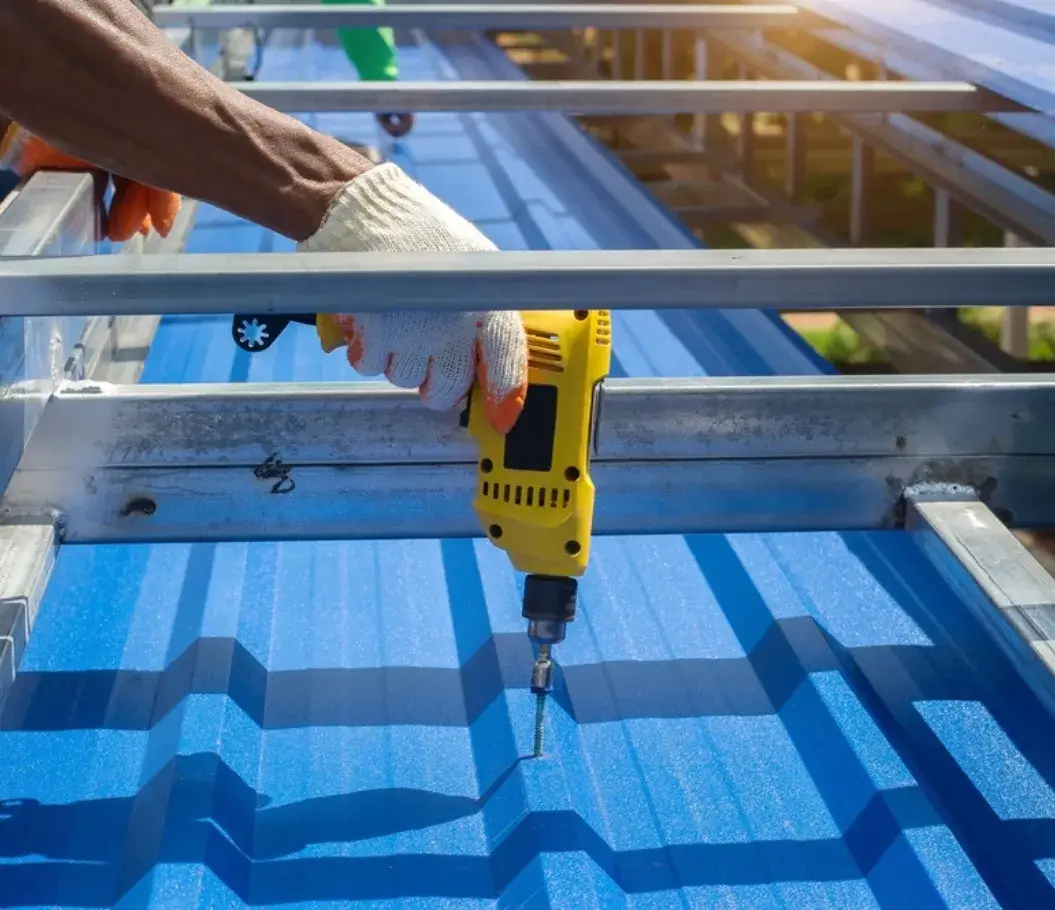 Worker installing a blue metal roof using a power drill.