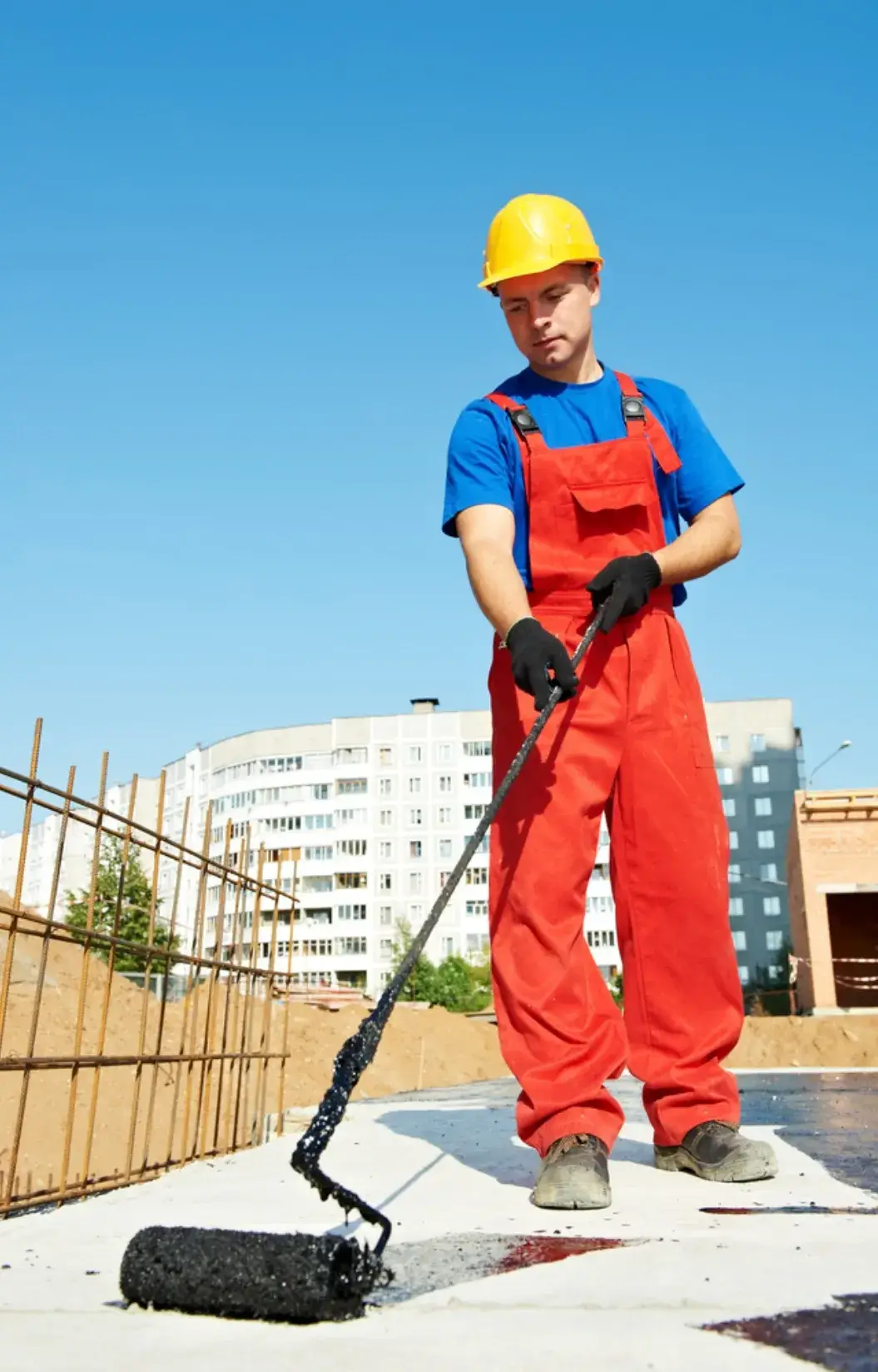 Construction worker applying waterproofing tar on a concrete surface with a roller.